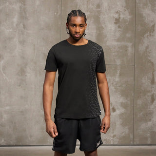 En mand står foran en grå betonvæg iført et sort træningssæt fra Everlast bestående af en t-shirt med diskret mønster over skulderen og sorte shorts med logo. Han har fletninger og et fokuseret udtryk, hvilket understreger det sporty og seriøse look.